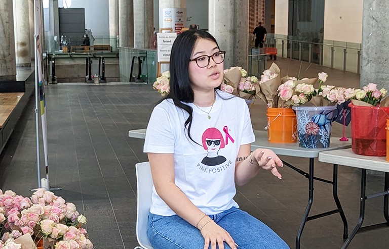 A young woman sits on a chair in a large interior space, speaking to people off-camera.