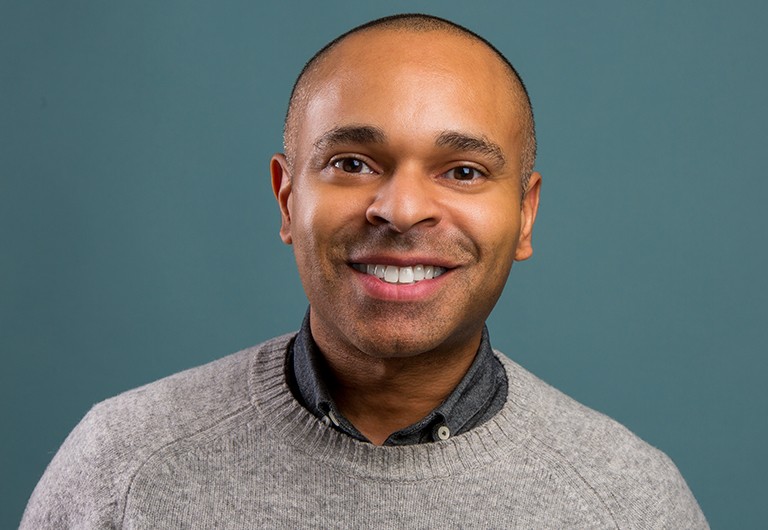 A smiling man with short, close-cropped hair, wearing a grey shirt and light grey sweater.