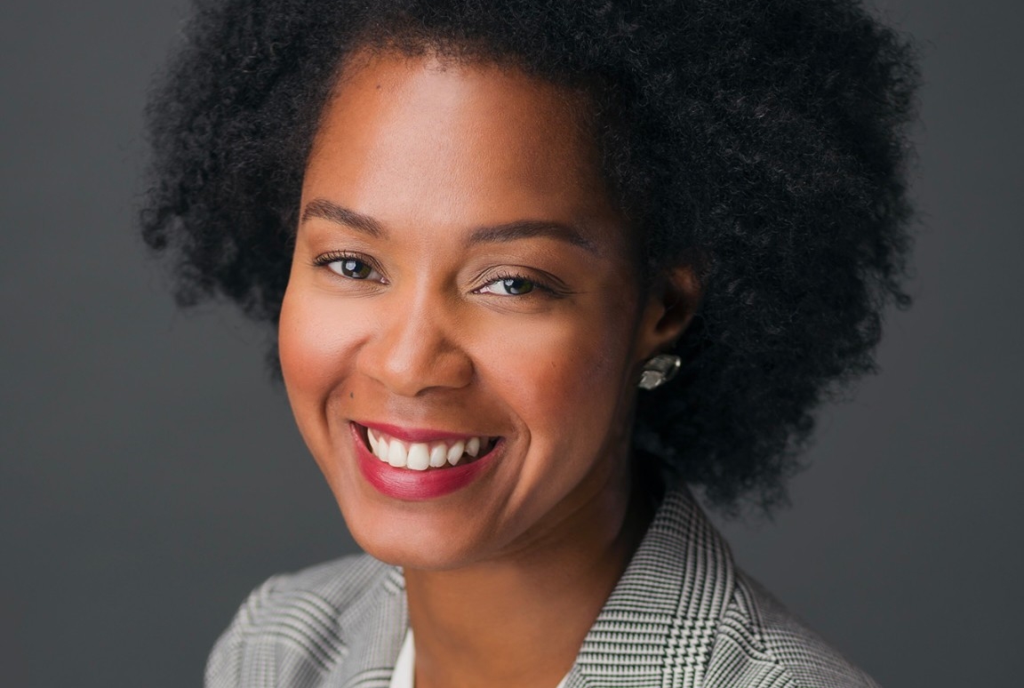 Smiling woman with curly, dark hair and wearing a white shirt and checkered blazer.