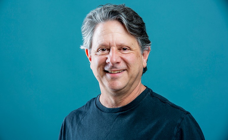 Smiling man in a blue T-shirt standing in front of a brick wall.