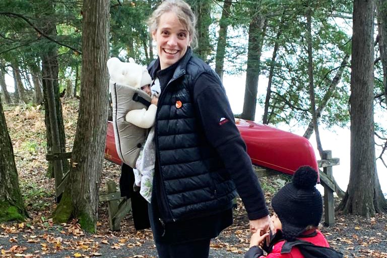 A smiling woman walking in the forest holding the hand of toddler and with a small baby in a sling across her front.