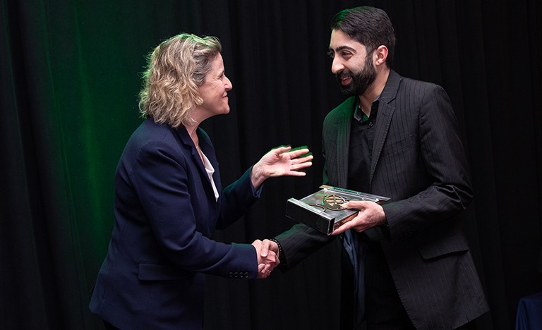 A woman with curly blonde hair giving an award and congratulations to a young man with dark hair and a beard, in a suit 