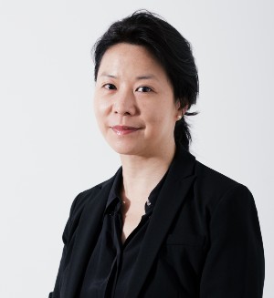 A woman with black hair wears dark clothing in front of a gray background