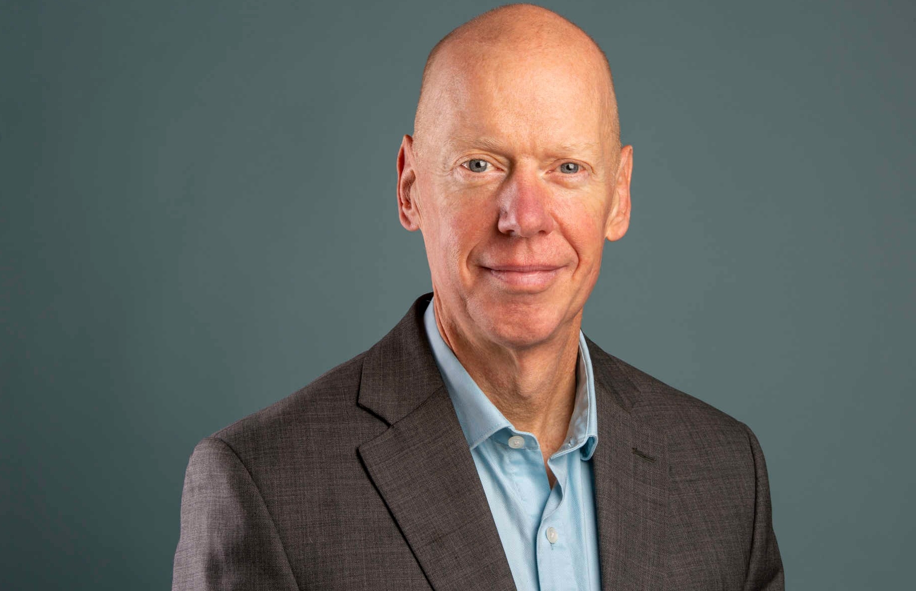 Smiling bald man, wearing a blue shirt and brown suit jacket.