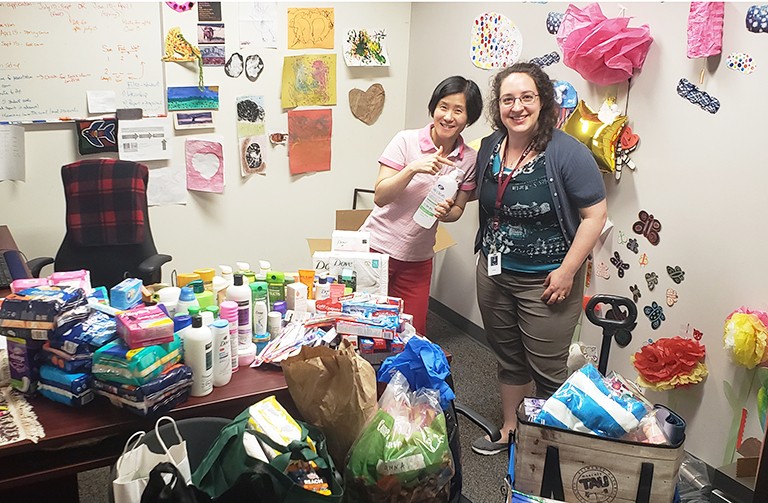 Two women standing in a office with piles of donated goods on the desk and chairs