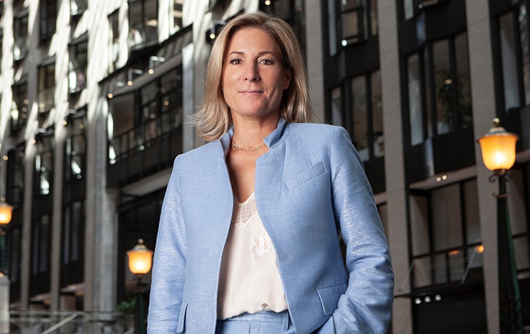 Smiling blonde woman wearing a light blue suit and pants with white shirt and with city buildings behind her.