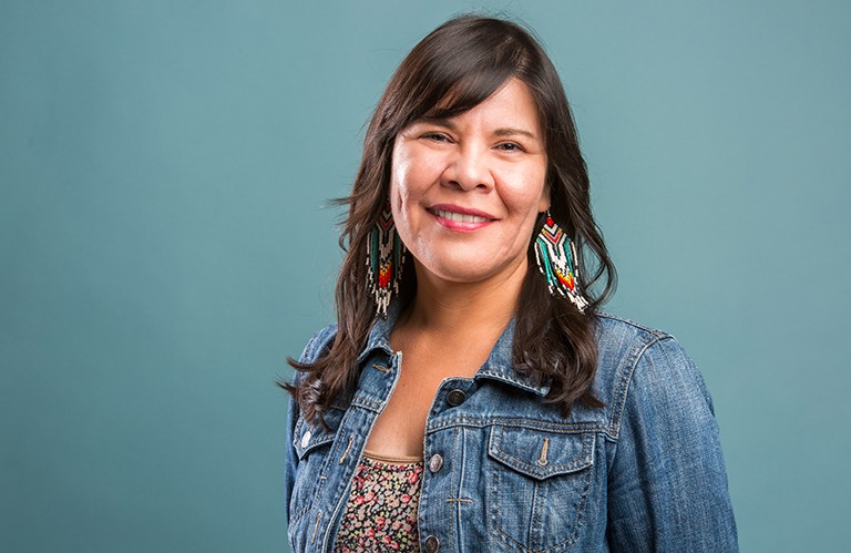 A smiling woman with long, dark hair, wearing a floral pattern top and a jean jacket