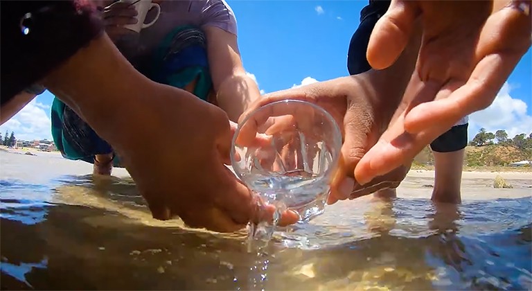 Image of several hands holding a glass with water spilling into the shallows of a river.