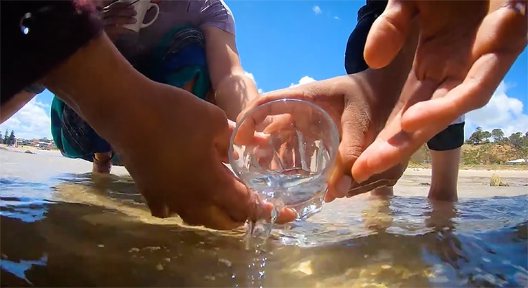 Video still from Franks’s documentary film, which centres around water and water-related concerns. Image of several hands holding a glass with water spilling into the shallows of a river.