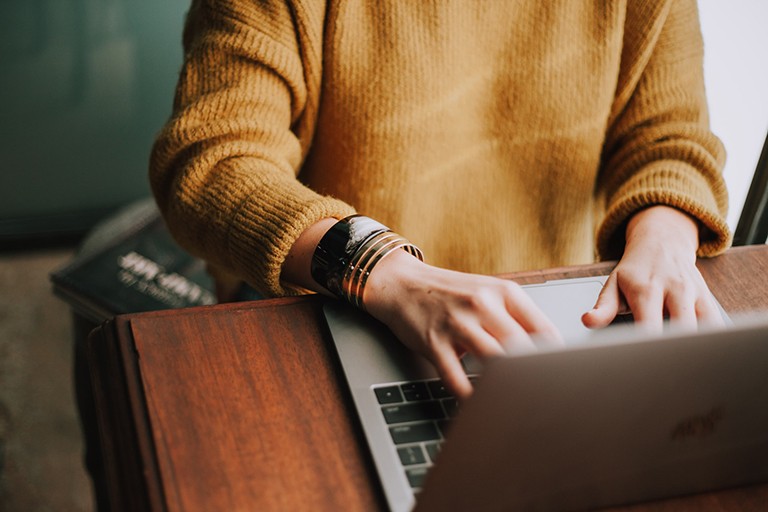 Someone seen from the chest down, using a laptop and wearing a mustard yellow sweater