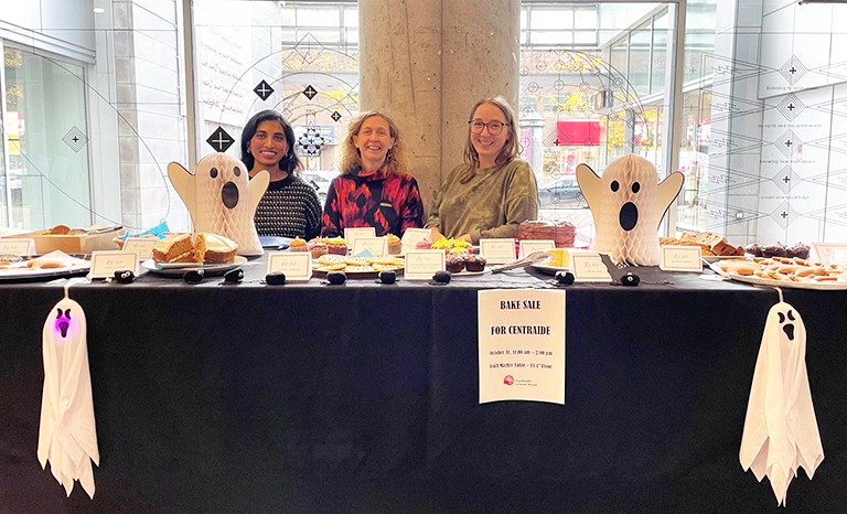 Three woman sitting at a table in an interior, with baked goods and halloween decorations on it.