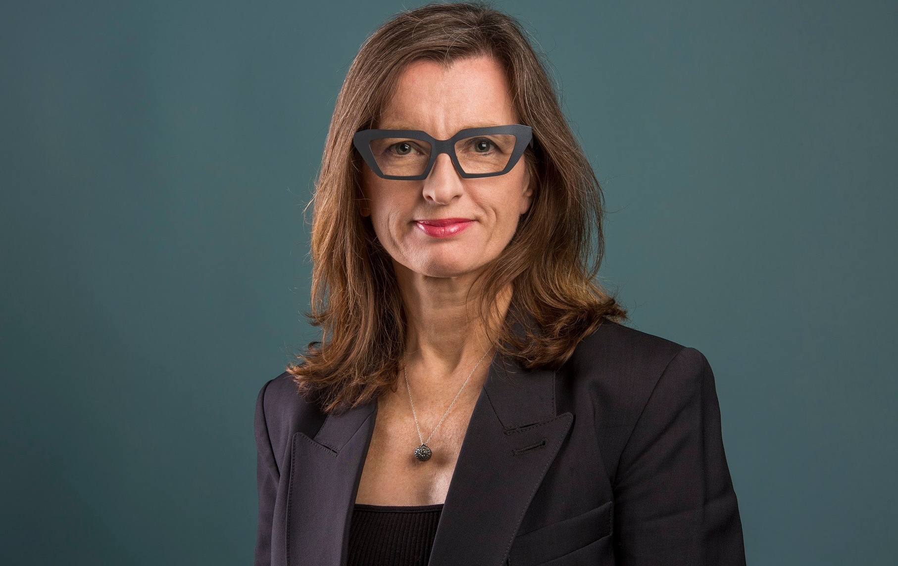 Smiling woman with long light-brown hair, wearing dark-rimmed glasses and a black top and blazer.