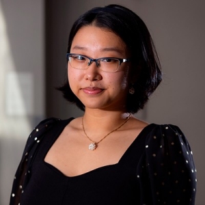 Smiling young woman with shoulder-length dark hair, glasses, and a black dress with small white polka dots.