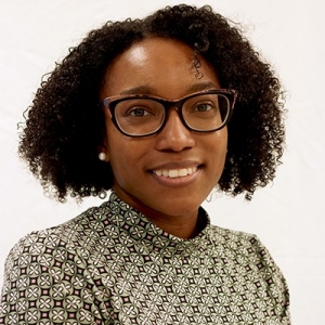 Smiling young woman with shoulder-length curly hair, dark-rimmed glasses, and wearing a pale patterned shirt.