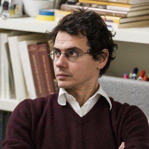 A person with glasses and curly hair is seated with folded arms, wearing a maroon sweater over a white shirt. Books and office supplies are visible in the background.