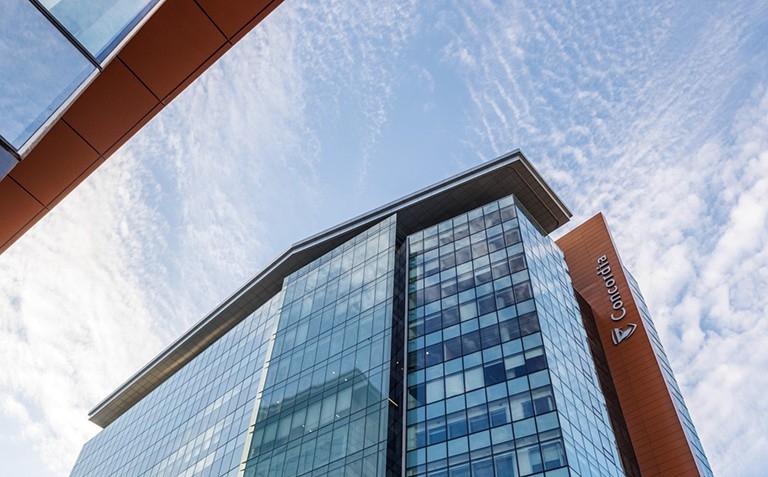 Quebec Superior Court rules in Concordia’s favour on tuition changes, francization People walk past a modern glass building with “CONCORDIA” written on the window, under a partly cloudy blue sky in an urban setting.