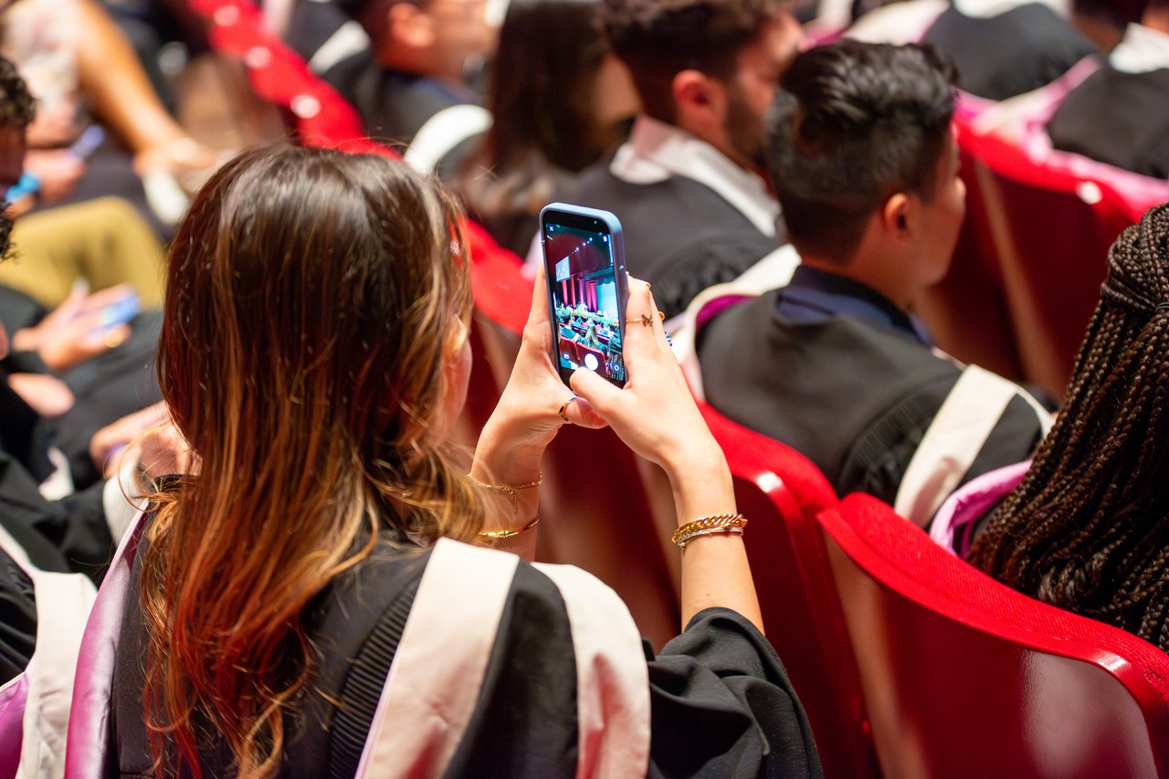 A young woman at a convocation ceremony seen from the back and holding a cellphone