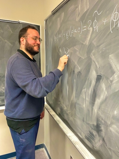 A man in a blue sweater with glasses writes math equations on a green chalkboard while smiling.