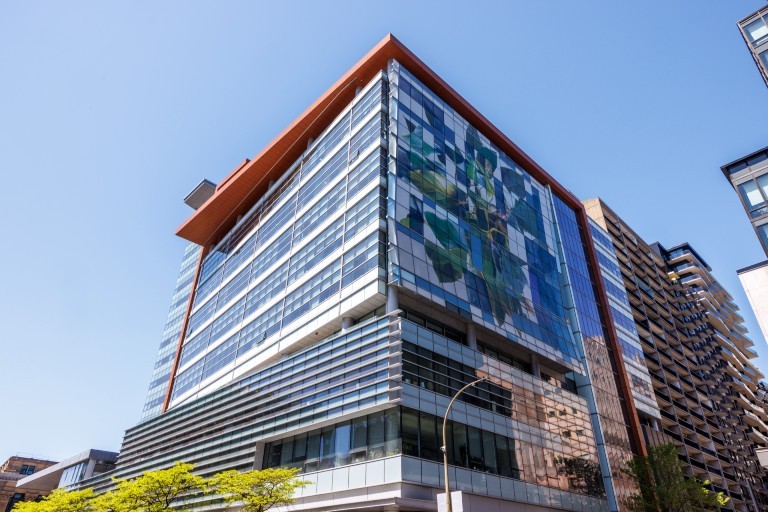 A modern glass office building with a colourful abstract mural on its facade, photographed from a low angle against a clear blue sky.