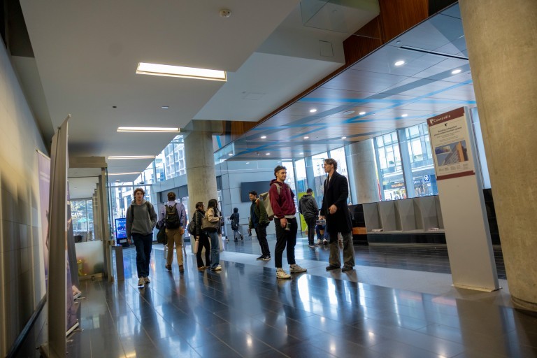 People walk and stand in a modern indoor hallway with tall windows, polished floors and informational signs.