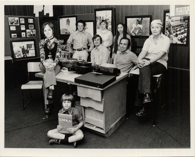 A black and white archival photo of a group of people sitting and standing around a desk and smiling for the camera.