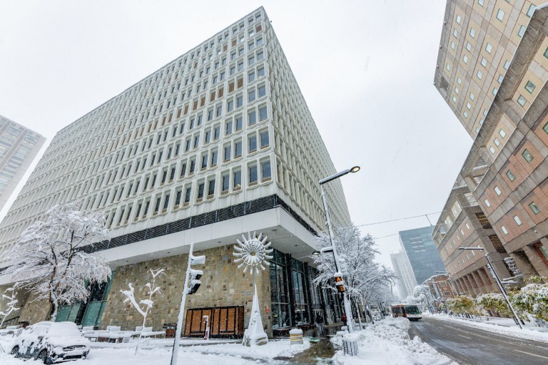 Winter view of the Henry F. Hall Building at the corner of De Maisonneuve Boulevard and Mackay Street, with snow-covered trees, sidewalks and pedestrians.