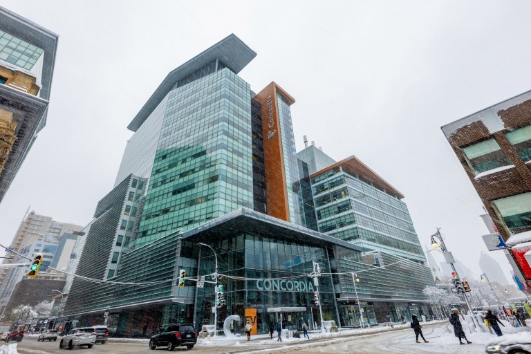 A winter street view of Concordia’s Sir George Williams Campus, with pedestrians walking and vehicles moving through the snowy intersection in front of the EV Building.