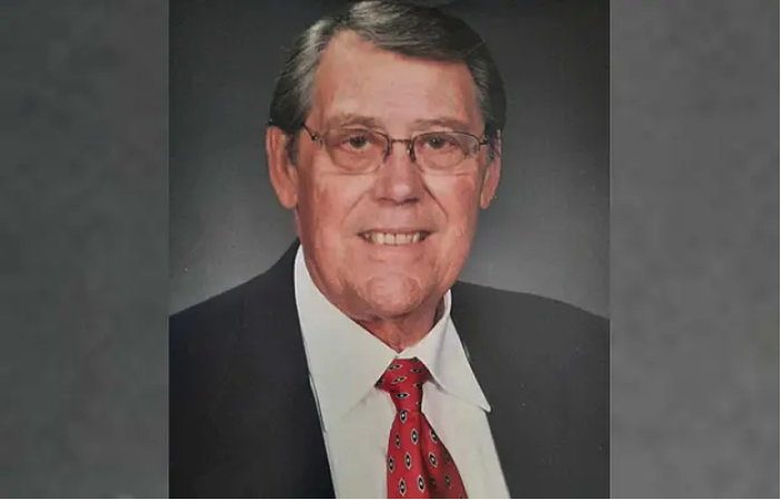 Studio portrait of an older man with short gray hair and glasses, wearing a dark suit jacket, white dress shirt, and red patterned tie against a neutral gray background.