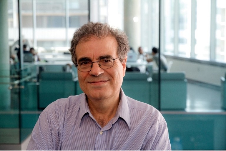 A smiling man wearing glasses and a pink polo, sitting in an office space