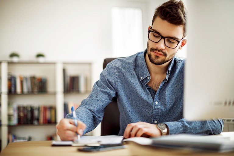 Young man with short dark hair and a beard and wearing dark-rimmed glasses, sitting at a desktop computer and writing on a paper on a clipboard.