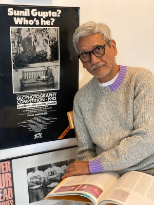 portrait of Sunil Gupta siting beside a poster from a 1983 exhibition of his work.