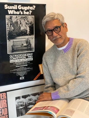 portrait of Sunil Gupta siting beside a poster from a 1983 exhibition of his work.