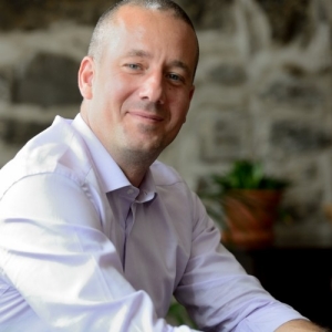 Mathieu Rondeau wears a white dress shirt, smiling, in front of an old stone wall