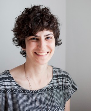 A smiling individual with curly dark hair who is wearing a short-sleeve blouse