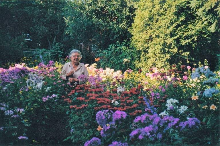 An elderly woman with white hair and glasses stands in the middle of a garden.