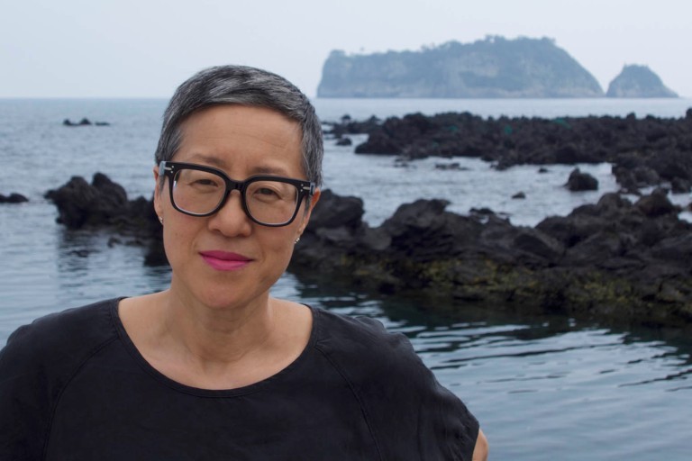 A woman with short hair and dark-rimmed glasses stands in front of a body of water.