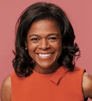 Woman with orange dress and short black hair smiles and poses in front of pink background.