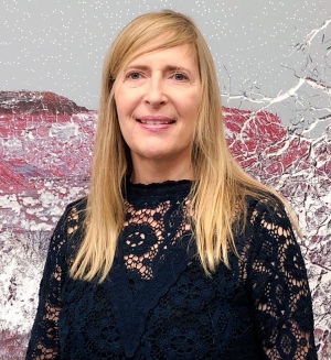 Woman with long blonde hair, wearing a black lace sweater, smiles as she stands in front of a pink, grey and white abstract painting.