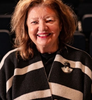Woman with brown hair wearing black and white striped wrap sweater stands and smiles in front of empty theatre seats.