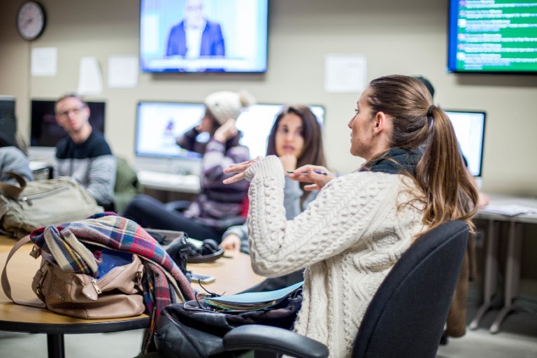 A student in a sweater sits at a desk talking to her colleagues in the background