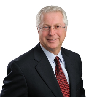 Richard Joly, wearing glasses, a dark suit and read tie, smiles warmly.