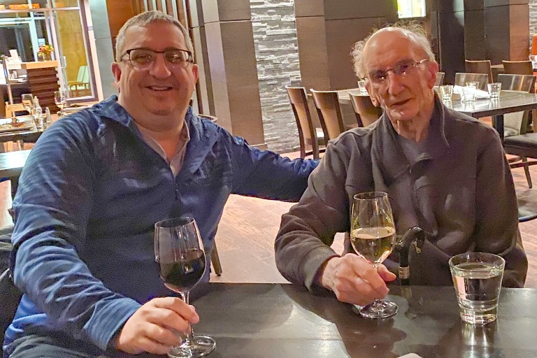 A father and son sit at a table while drinking glasses of white and red wine.