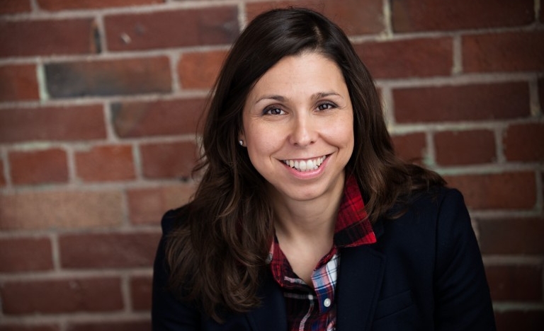 A woman wearing a jacket and flannel button-up shirt sits in front of a brick wall.