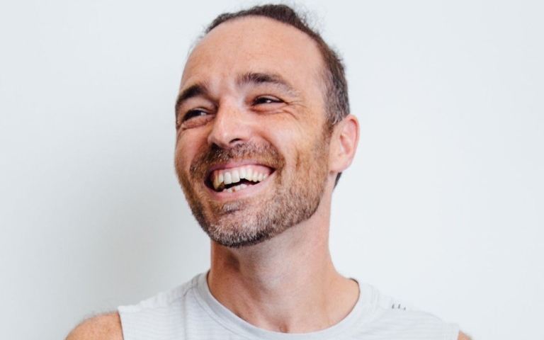 A man in a white tank top laughs and looks to his right.