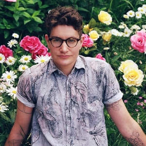 A man wearing glasses and with tattoos on his arms stands in front of flowers.