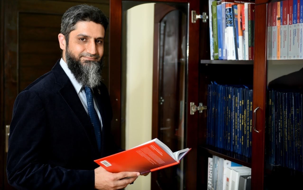 Man with dark hair and beard wearing a suit and tie stands in a library, holding open a red book