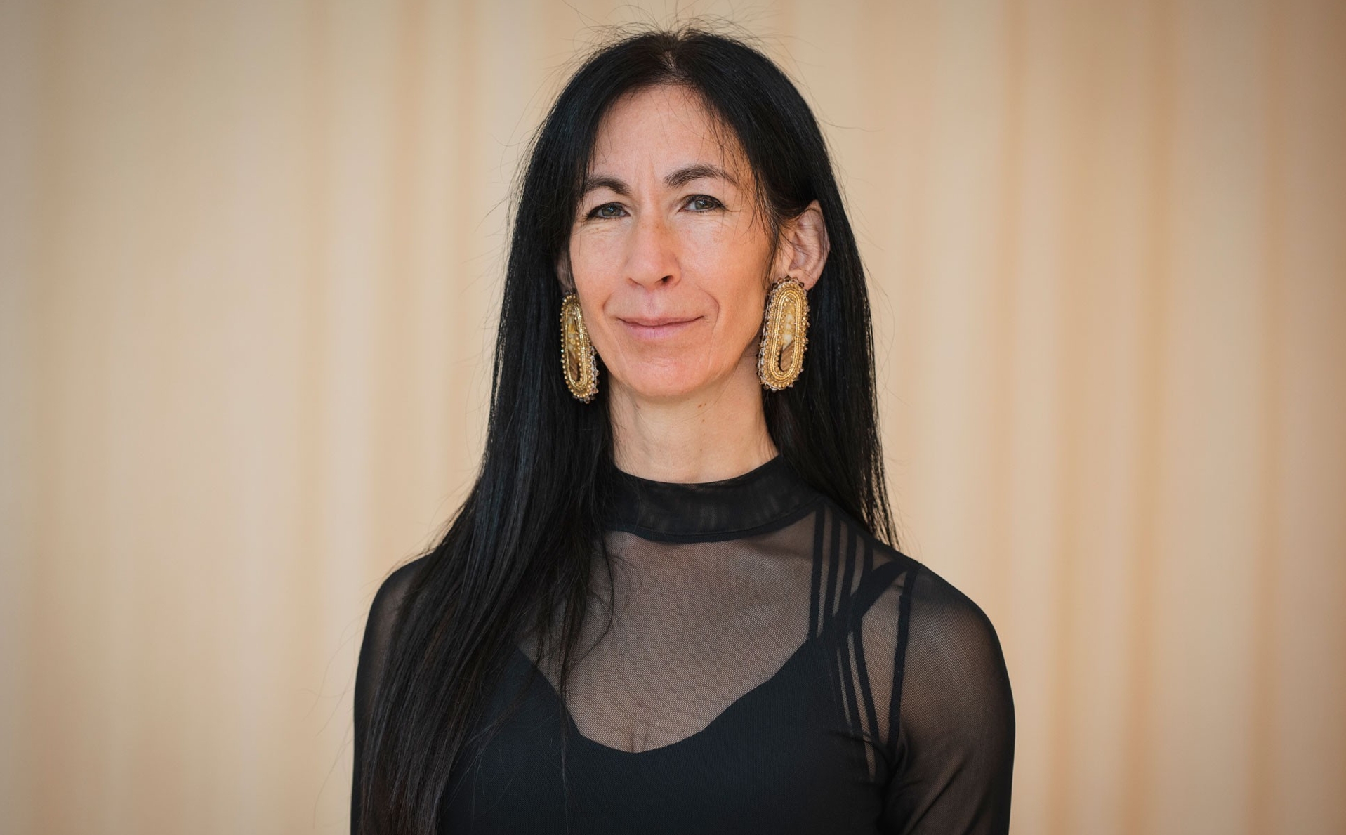 Indigenous woman with black hair wears gold earrings and black blouse, stands in front of beige background