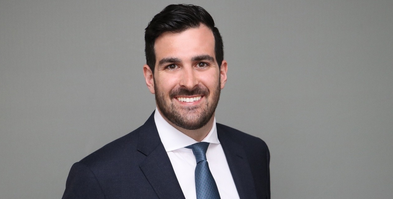 Alex Leopold, wearing a dark suit and tie, smiles warmly