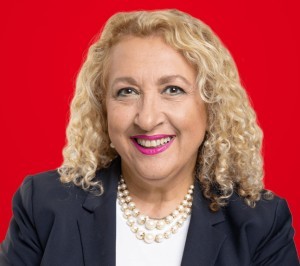 Headshot of a woman with blonde curly hair wearing a pearl necklace and suit jacket, red background