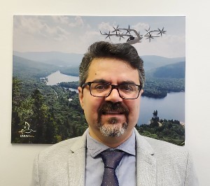 A man with dark hair, glasses and a goatee stands in front of landscape poster with a helicopter